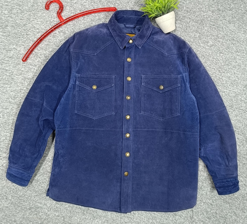 Peut inclure: Une chemise en velours c&ocirc;tel&eacute; bleu marine avec deux poches de poitrine et des boutons dor&eacute;s. La chemise a un col classique et des manches longues. La chemise est pr&eacute;sent&eacute;e sur un cintre rouge.