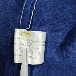 Peut inclure: Gros plan sur un tissu bleu fonc&eacute; avec une &eacute;tiquette blanche attach&eacute;e. L'&eacute;tiquette contient du texte dont "NO. 6757806" et "100% POLYESTER". Des ciseaux coupent l'&eacute;tiquette. Le tissu a un aspect doux et textur&eacute;.