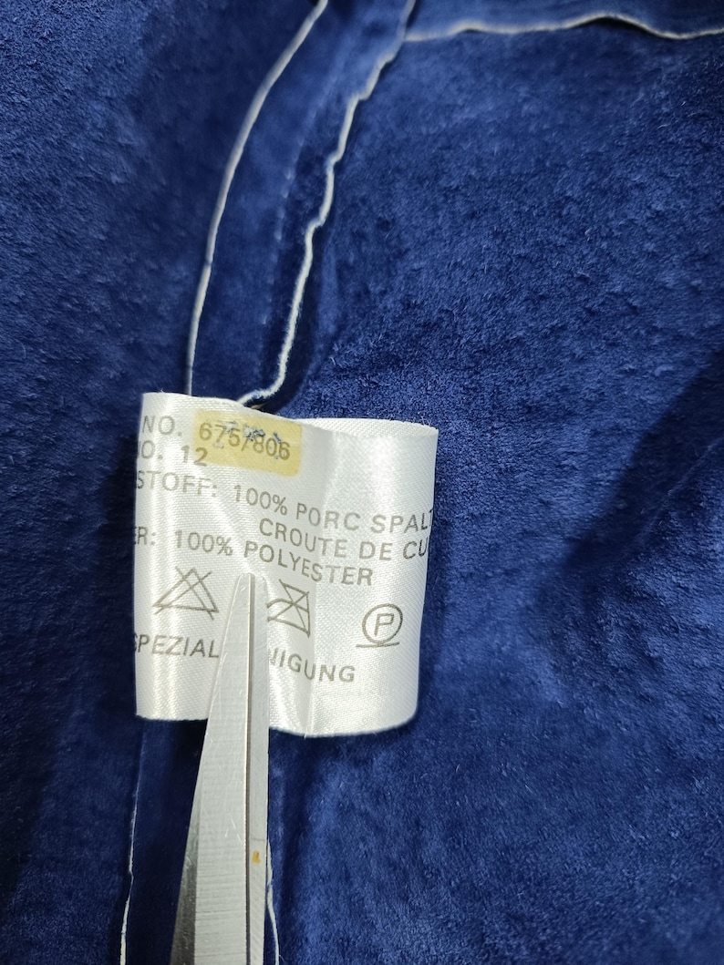 Peut inclure: Gros plan sur un tissu bleu fonc&eacute; avec une &eacute;tiquette blanche attach&eacute;e. L'&eacute;tiquette contient du texte dont "NO. 6757806" et "100% POLYESTER". Des ciseaux coupent l'&eacute;tiquette. Le tissu a un aspect doux et textur&eacute;.