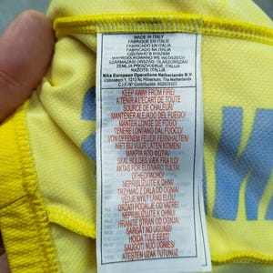 Puede incluir: Primer plano de una tela amarilla con una etiqueta. La etiqueta contiene texto en varios idiomas, incluyendo "MADE IN ITALY" e instrucciones de seguridad contra incendios. La tela tiene un patr&oacute;n sutil y un dise&ntilde;o azul.