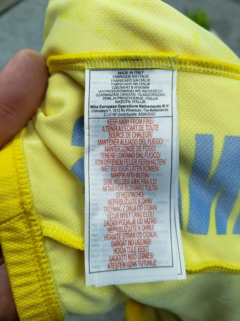 Puede incluir: Primer plano de una tela amarilla con una etiqueta. La etiqueta contiene texto en varios idiomas, incluyendo "MADE IN ITALY" e instrucciones de seguridad contra incendios. La tela tiene un patr&oacute;n sutil y un dise&ntilde;o azul.