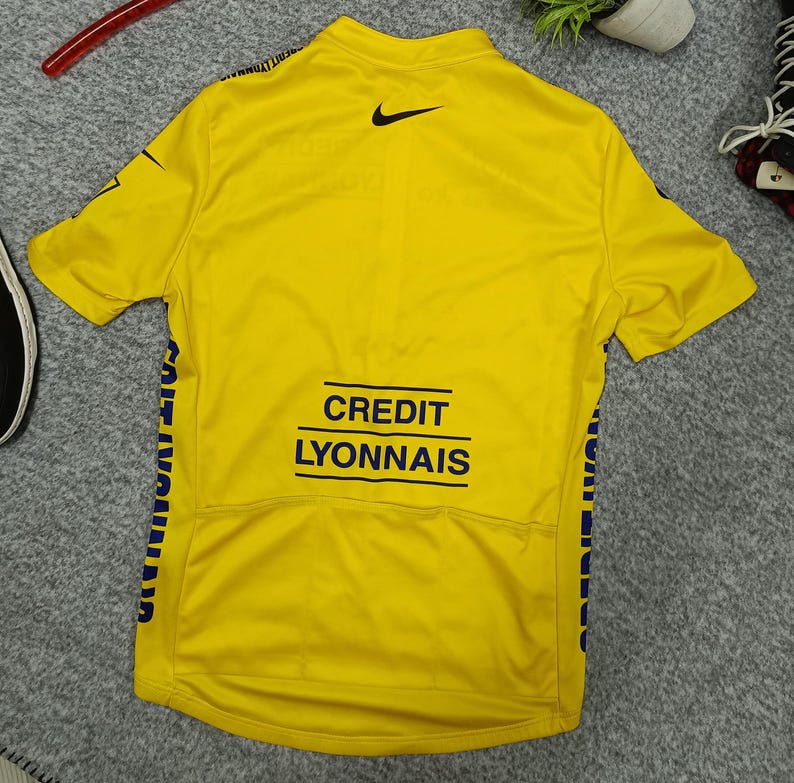 Puede incluir: Maillot de ciclismo amarillo con el logo de Nike y las palabras "CREDIT LYONNAIS" en negro. El maillot tiene detalles azules y texto en las mangas. El maillot es de manga corta y tiene cuello.