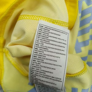 Puede incluir: Primer plano de una camiseta deportiva amarilla con un estampado azul claro en la manga. Se ve una etiqueta con texto en varios idiomas, que indica que el tejido es 100% poli&eacute;ster, sin incluir la decoraci&oacute;n.