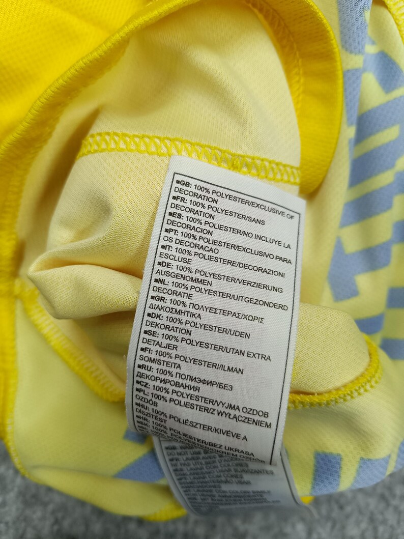 Puede incluir: Primer plano de una camiseta deportiva amarilla con un estampado azul claro en la manga. Se ve una etiqueta con texto en varios idiomas, que indica que el tejido es 100% poli&eacute;ster, sin incluir la decoraci&oacute;n.