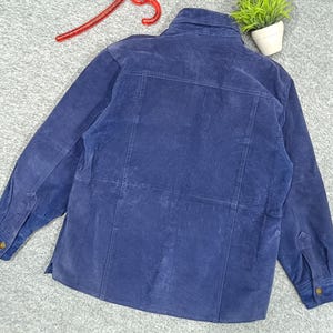 Peut inclure: Veste en daim bleu marine avec fermeture boutonn&eacute;e, manches longues et col. La veste a un design classique avec une coupe ajust&eacute;e et comporte des poignets boutonn&eacute;s et une poche poitrine. Le dos de la veste est visible, montrant les coutures et la texture du daim.