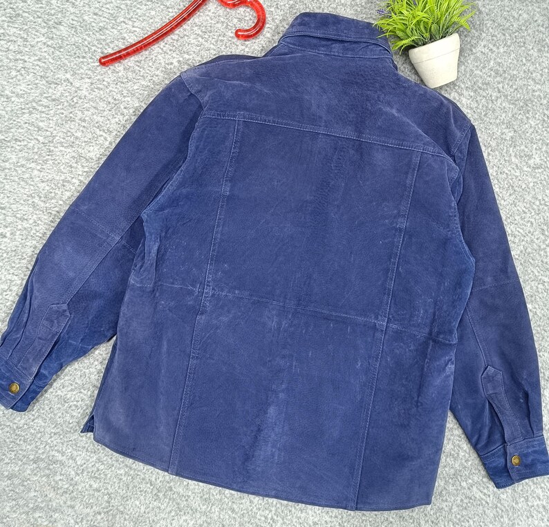 Peut inclure: Veste en daim bleu marine avec fermeture boutonn&eacute;e, manches longues et col. La veste a un design classique avec une coupe ajust&eacute;e et comporte des poignets boutonn&eacute;s et une poche poitrine. Le dos de la veste est visible, montrant les coutures et la texture du daim.