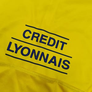 Puede incluir: Primer plano de una tela amarilla con el texto azul "CREDIT LYONNAIS". El texto, en negrita y sin serifa, est&aacute; subrayado por dos l&iacute;neas paralelas. La tela parece texturizada.