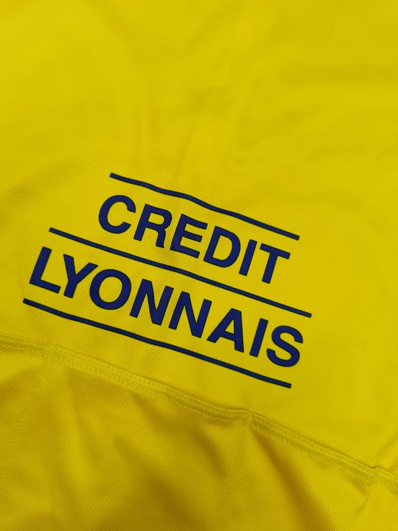 Puede incluir: Primer plano de una tela amarilla con el texto azul "CREDIT LYONNAIS". El texto, en negrita y sin serifa, est&aacute; subrayado por dos l&iacute;neas paralelas. La tela parece texturizada.