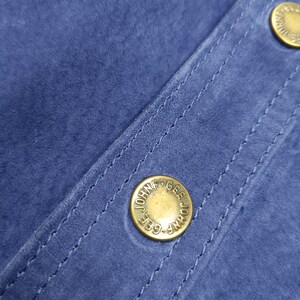 Peut inclure: Gros plan sur un tissu en daim bleu avec des coutures visibles et deux boutons dor&eacute;s. Les boutons portent l'inscription "JOHN GEE". Le tissu a une texture douce et velout&eacute;e.