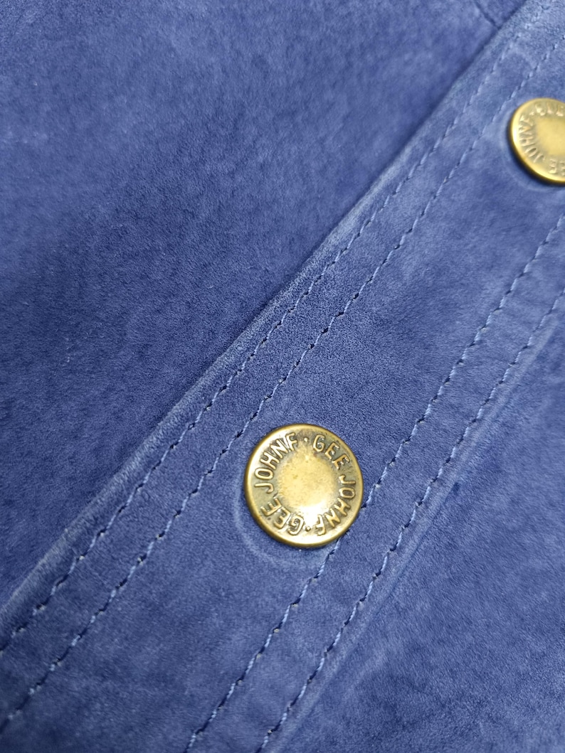 Peut inclure: Gros plan sur un tissu en daim bleu avec des coutures visibles et deux boutons dor&eacute;s. Les boutons portent l'inscription "JOHN GEE". Le tissu a une texture douce et velout&eacute;e.