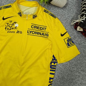 Puede incluir: Maillot de ciclismo amarillo con el logo de Nike en negro y texto azul que dice "CREDIT LYONNAIS" y "le TOUR de FRANCE 2005". El maillot tiene cremallera y mangas cortas. El maillot es de color amarillo brillante.