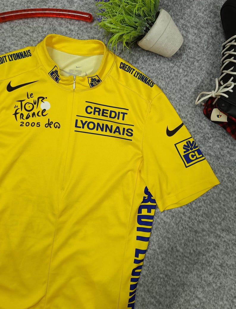 Puede incluir: Maillot de ciclismo amarillo con el logo de Nike en negro y texto azul que dice "CREDIT LYONNAIS" y "le TOUR de FRANCE 2005". El maillot tiene cremallera y mangas cortas. El maillot es de color amarillo brillante.