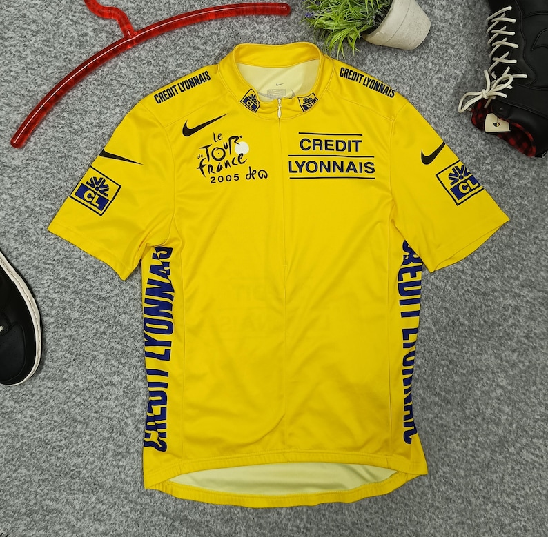 Puede incluir: Un maillot de ciclismo amarillo con logotipos negros de Nike y el texto "CREDIT LYONNAIS" y "le TOUR de France 2005". El maillot tiene cremallera y mangas cortas. El maillot tiene el logo "CL" en las mangas.