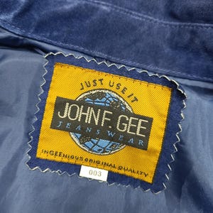 Peut inclure: Gros plan sur une veste en daim bleu avec une &eacute;tiquette jaune. L'&eacute;tiquette indique "JOHN F. GEE JEANSWEAR" avec le texte "JUST USE IT" au-dessus et "INGENIOUS ORIGINAL QUALITY" en dessous. Le num&eacute;ro "003" est imprim&eacute; sur une &eacute;tiquette blanche.
