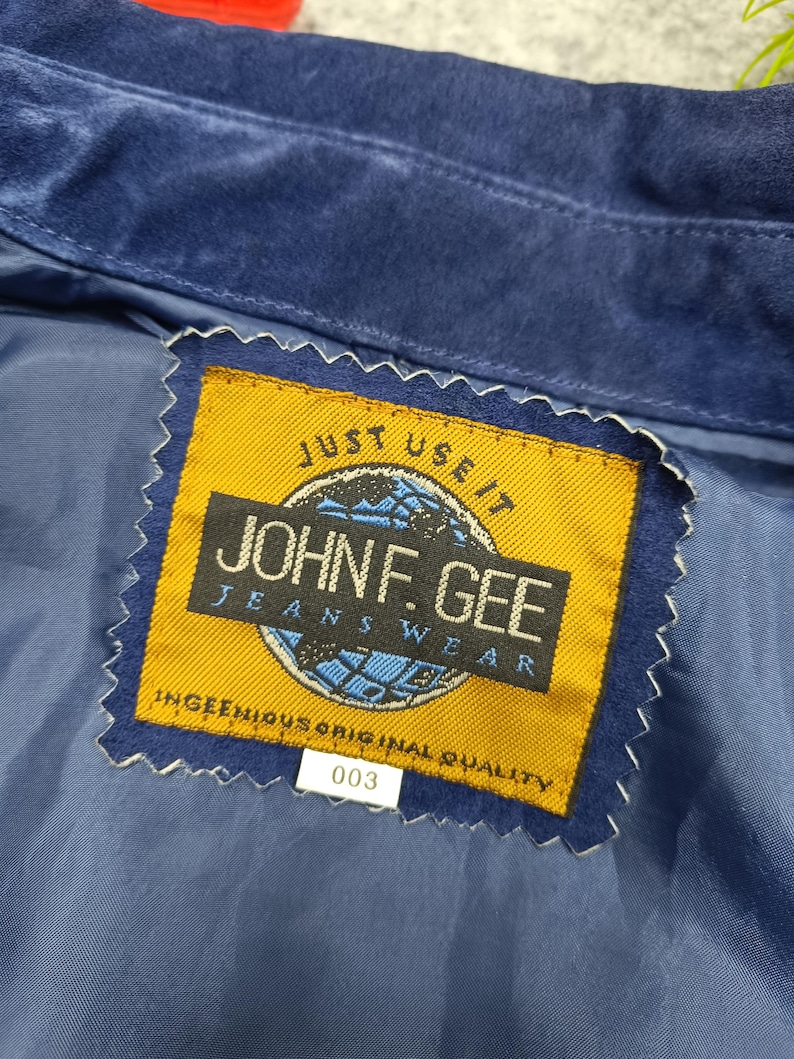 Peut inclure: Gros plan sur une veste en daim bleu avec une &eacute;tiquette jaune. L'&eacute;tiquette indique "JOHN F. GEE JEANSWEAR" avec le texte "JUST USE IT" au-dessus et "INGENIOUS ORIGINAL QUALITY" en dessous. Le num&eacute;ro "003" est imprim&eacute; sur une &eacute;tiquette blanche.