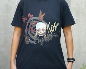 Peut inclure: Un t-shirt noir à manches courtes avec une impression graphique portant le texte See You On The Other Side et Korn. Le motif comprend un visage pâle aux yeux écarquillés, les mains sur la bouche, une silhouette de lapin et une spirale rouge et blanche.