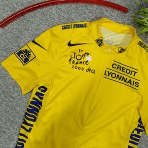Puede incluir: Maillot de ciclismo amarillo con el logo de Nike negro y texto "le Tour de France 2005". Presenta "CREDIT LYONNAIS" y otros logotipos en negro y azul. Manga corta, con cremallera parcial y una percha roja.