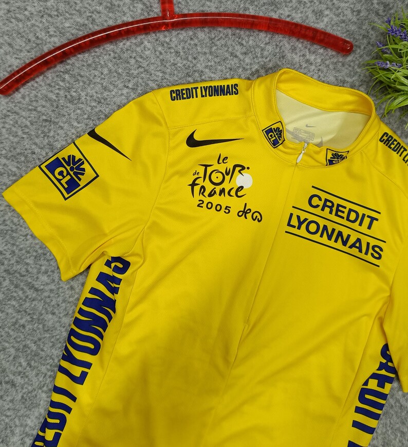 Puede incluir: Maillot de ciclismo amarillo con el logo de Nike negro y texto "le Tour de France 2005". Presenta "CREDIT LYONNAIS" y otros logotipos en negro y azul. Manga corta, con cremallera parcial y una percha roja.