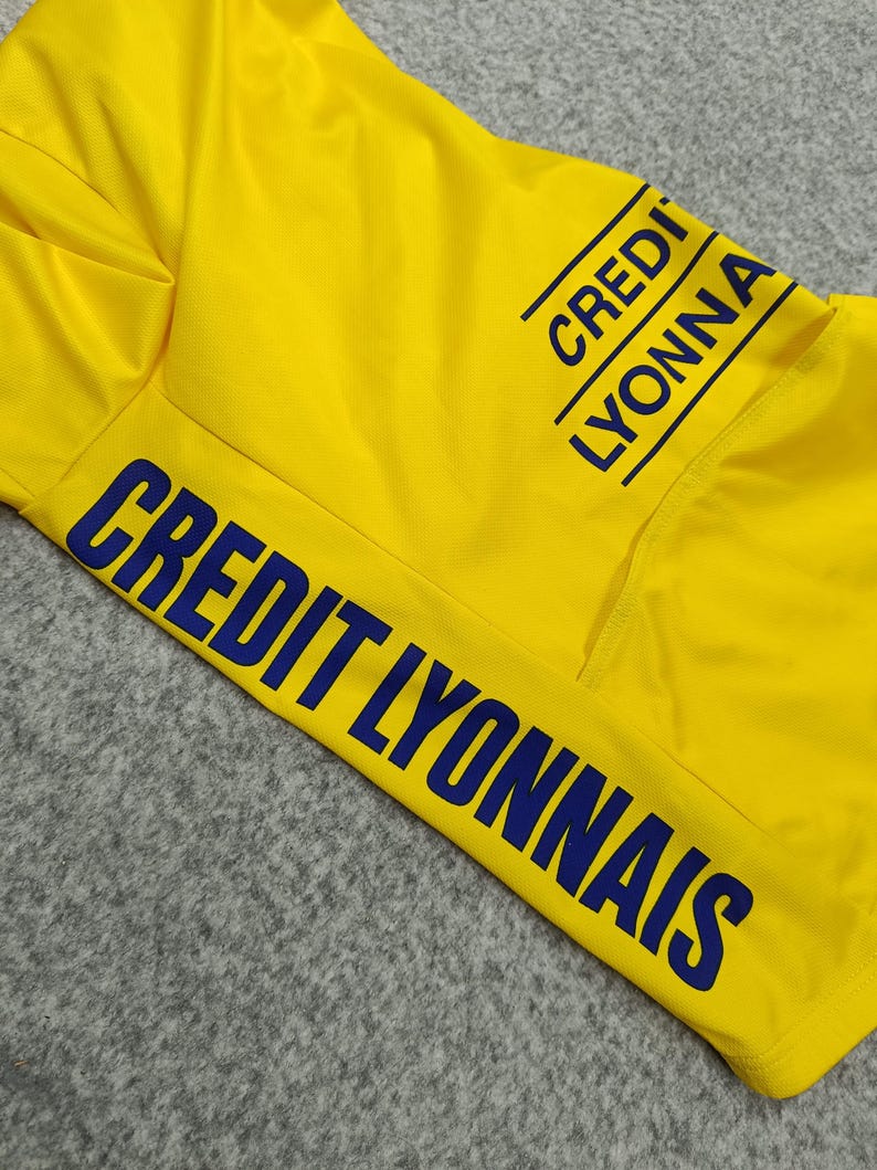 Puede incluir: Camiseta deportiva amarilla brillante con la inscripci&oacute;n "CREDIT LYONNAIS" en azul marino. La camiseta tiene un bolsillo y el texto se repite en la manga. La tela parece ser un material ligero y transpirable.
