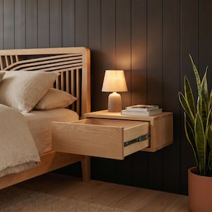 May include: A light-colored wooden floating nightstand with an open drawer featuring visible metal glides. A small lamp with a beige shade and a cylindrical base sits on top of the nightstand next to a stack of two books.
