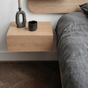 May include: A light oak floating nightstand mounted to a white wall. On the surface sits a dark gray textured sculptural candle holder with a thin taper candle and a small, matte black cylindrical cup. A dark gray linen bed is positioned nearby.