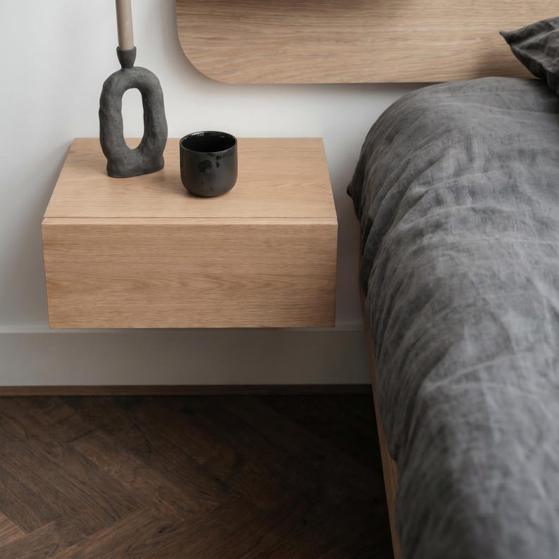 May include: A light oak floating nightstand mounted to a white wall. On the surface sits a dark gray textured sculptural candle holder with a thin taper candle and a small, matte black cylindrical cup. A dark gray linen bed is positioned nearby.