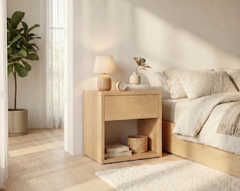 May include: A light-toned wooden nightstand featuring a single top drawer and an open lower shelf. The nightstand is topped with a small lamp with a beige shade, a round clock, and a small vase with dried stems. A woven basket sits on the shelf.