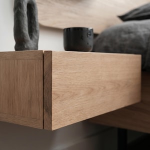 May include: A light brown oak wood floating nightstand with a rectangular drawer front. On top of the nightstand sits a black textured sculptural object and a small black ceramic cup. The wood grain is visible across the horizontal surface.