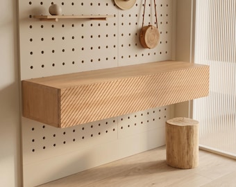 Puede incluir: Una consola flotante de madera clara con un patrón de ranuras diagonales en el frente, montada en una pared de tablero perforado beige. El conjunto incluye un estante pequeño con una esfera, un sombrero de paja, un bolso redondo y un taburete.