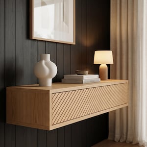 Peut inclure: Une étagère murale flottante en chêne clair avec une façade de tiroir à lattes diagonales texturées. Sur l'étagère se trouvent un vase bulbeux blanc mat, un petit bol gris et deux livres empilés. Une lampe de table en bois avec un abat-jour blanc.