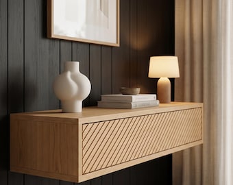 Könnte beinhalten: Ein schwebendes Wandregal aus heller Eiche mit einer strukturierten Schubladenfront aus diagonalen Lamellen. Auf dem Regal stehen eine matte, bauchige weiße Vase, eine kleine graue Schale und zwei gestapelte Bücher. Eine kleine Holztischlampe mit weißem Schirm.