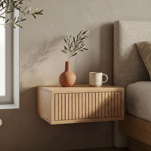 Könnte beinhalten: Ein schwebender Nachttisch aus hellem Holz mit einer einzelnen Schublade mit vertikalen Lamellen an der Vorderseite. Eine matte, terrakottafarbene runde Vase mit einem belaubten Zweig und eine weiße Keramiktasse mit Marmormuster stehen darauf.