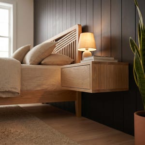 May include: A light wood floating nightstand with a single drawer, mounted to a dark paneled wall next to a bed. The nightstand features a smooth, natural wood grain finish and supports a small lamp and a stack of books on its flat top surface.