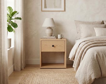 May include: A light oak nightstand featuring a single drawer with a small black circular knob and an open storage shelf underneath. The nightstand is positioned beside a bed with beige bedding and holds a table lamp with a white shade.