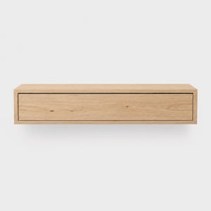 Könnte beinhalten: Ein schwebender, rechteckiger Holzschubkasten in hellem Eichenholz. Die Schublade hat ein schlichtes, modernes Design mit einer einzigen, zurückgesetzten Schubladenfront. Die Holzmaserung ist sichtbar und verleiht Textur. Für minimalistische Einrichtung.