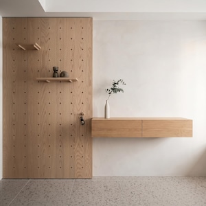 Può includere: Un pannello a parete forato in legno chiaro con file verticali di fori e due piccoli ripiani regolabili. Accanto, una console sospesa in legno chiaro con due cassetti sostiene un vaso bianco con un rametto verde. Una chiave è appesa al pannello.