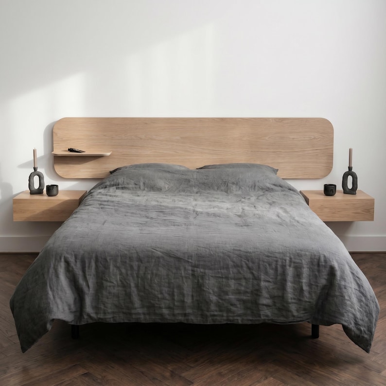 May include: A light oak wood headboard with rounded corners and an integrated floating shelf, paired with two matching floating nightstands. The bed is dressed in a dark gray linen duvet cover, and each nightstand holds a dark gray candle holder.