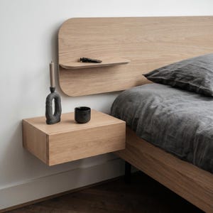 Peut inclure: Une table de chevet flottante en bois de chêne clair fixée à une tête de lit assortie aux bords arrondis. La table de chevet supporte un bougeoir texturé gris foncé avec une bougie et un petit bol noir. Un oreiller en lin gris foncé est posé.