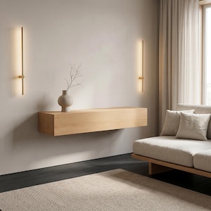 Può includere: Una mensola sospesa in legno chiaro montata a parete, con un vaso in ceramica opaca contenente un ramo sottile. Due applique verticali e sottili color oro con illuminazione a LED lineare integrata sono posizionate ai lati della mensola.