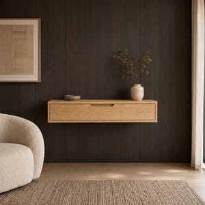 Puede incluir: Un estante flotante de madera clara con un cajón horizontal único y tirador empotrado. El estante está montado en una pared de paneles de madera oscura y sostiene un pequeño cuenco gris y un jarrón de cerámica beige con ramas verdes.