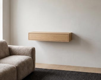 May include: A rectangular, light-toned wooden floating shelf featuring a single horizontal drawer. The unit is mounted directly onto a plain wall, providing a clean, linear storage solution with a smooth, natural wood grain finish and no visible hardware.