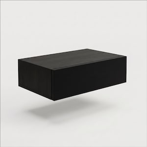 May include: A rectangular floating wall shelf with a dark black wood grain finish. The unit features a clean, boxy design with a flat top surface and a solid front panel, presenting a simple, functional storage solution for mounting on a wall.