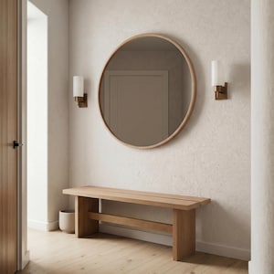 May include: A large circular wall mirror with a light wood frame, positioned above a rectangular wooden bench. Two wall-mounted sconces with white cylindrical shades flank the mirror, providing illumination on the textured off-white wall.