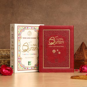 May include: A red leather-bound copy of The Holy Quran with gold lettering and embossed floral designs. Beside it is a matching box with the same title and design. The box is cream-coloured with colourful floral accents.