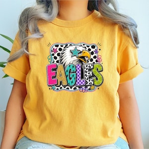May include: Mustard yellow t-shirt featuring a colorful graphic of an eagle with a star and the word "EAGLES" in a playful font. The design includes a black and white polka dot pattern and smiley faces.