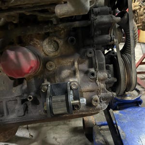 May include: A dark gray metal engine mount bracket with a cylindrical bushing and a threaded bolt, installed on the side of a weathered, grime-covered automotive engine block featuring a red oil filter and a black rubber drive belt.