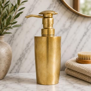 Può includere: Un dispenser di sapone cilindrico con finitura oro metallizzato e meccanismo a pompa abbinato. Il dispenser presenta una superficie liscia leggermente strutturata e un corpo affusolato, posizionato verticalmente come accessorio da bagno.
