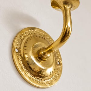 Op de afbeelding: Een gepolijste messing wandhaak met een ronde montageplaat voorzien van een decoratieve geschulpte rand en radiale gegraveerde lijnen. De haak heeft een gebogen arm die eindigt in een plat, rond uiteinde voor ophangdoeleinden.