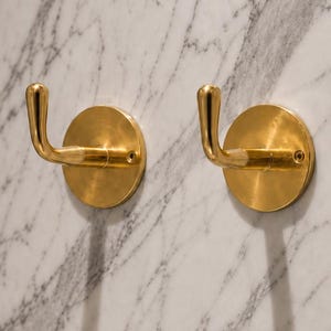 Op de afbeelding: Twee bijpassende wandhaken van gepolijst messing. Elke haak heeft een ronde basisplaat met een zichtbaar schroefgat en een gebogen, omhoog gerichte arm met een afgeronde punt, ontworpen om voorwerpen stevig aan een vlakke muur te hangen.