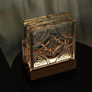 Op de afbeelding: Een decoratieve glazen bloklamp met een ingewikkeld patroon, van binnenuit verlicht. De lamp staat op een donkere houten voet, die een warme gloed afgeeft. De lamp staat op een zwarte tafel.